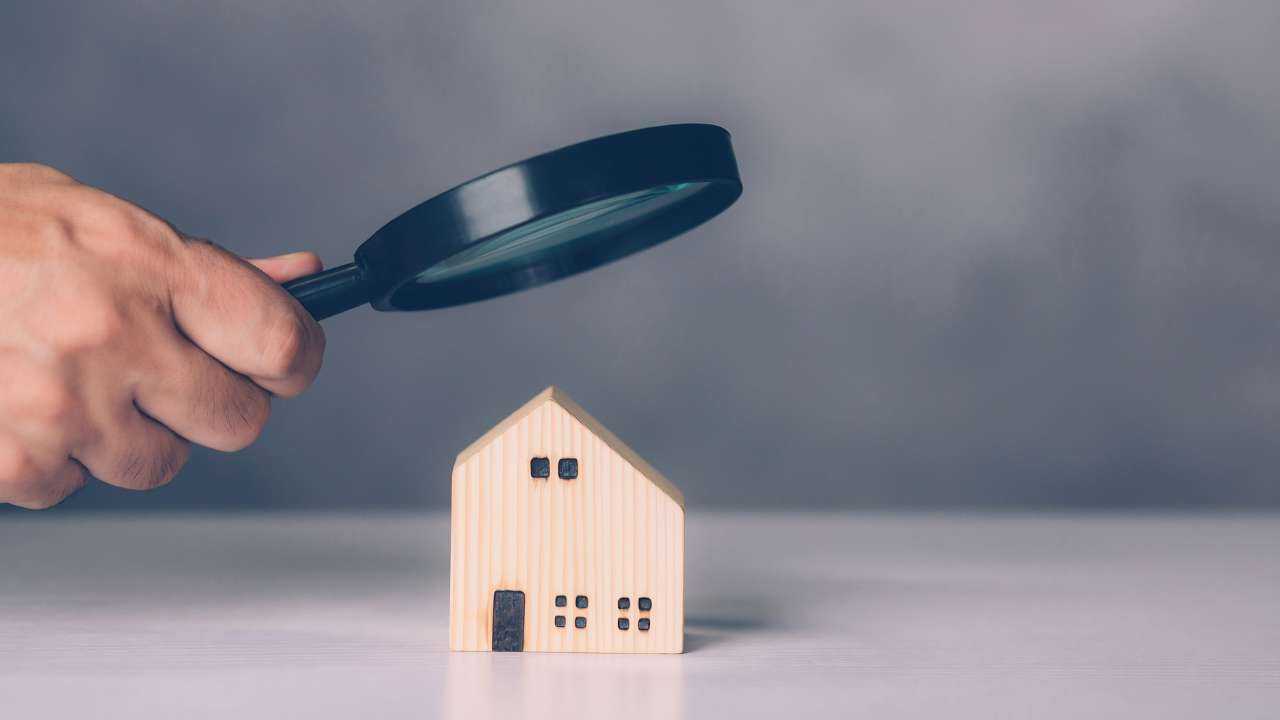 Деревяный макет домика, который под лупой разглядывает владелец, которому надо оплатить налог на недвижимое имущество
