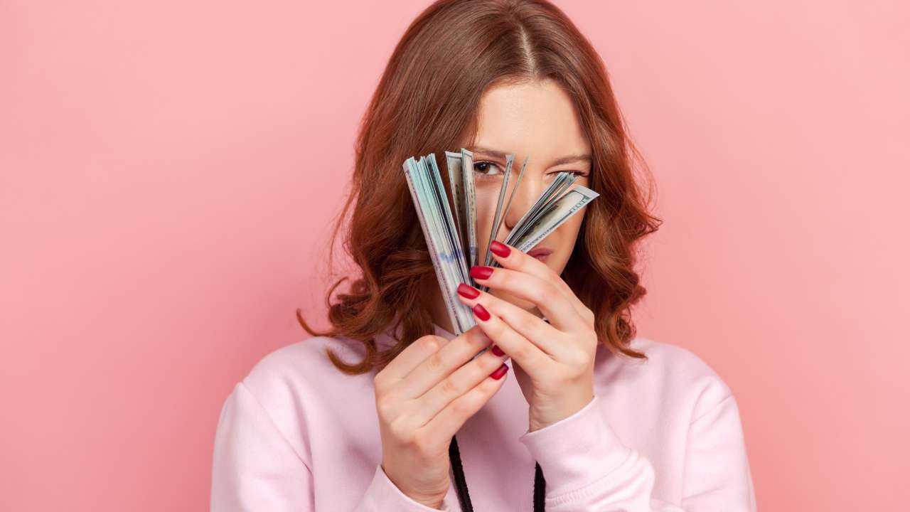 Девушка смотрит одним глазом через пачку купюр, которые получила благодаря пассивному доходу в интернете